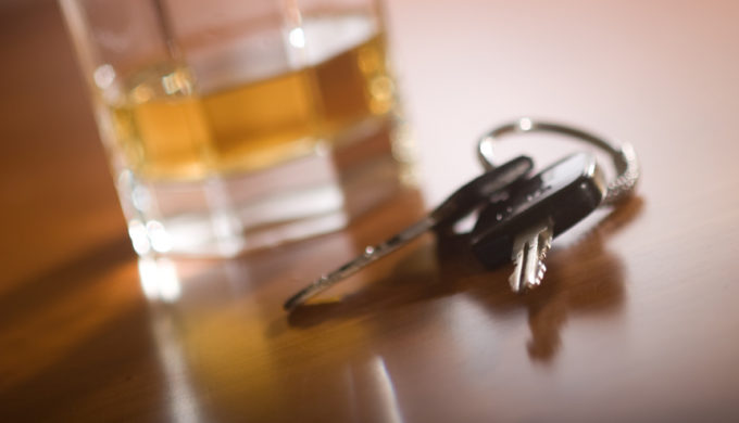 A set of car keys in the foreground and glass of whiskey behind.